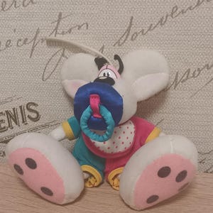 May include: A plush mouse toy with large white ears, wearing a multi-colored outfit with pink, teal, and yellow accents. The mouse has a white bib with red polka dots and a blue pacifier with a teal ring attached to its mouth. The feet are pink.