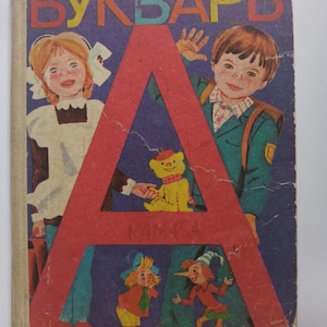 Op de afbeelding: Een vintage hardcover boek met de titel BUKVAR in grote kleurrijke letters bovenaan. De kaft toont een grote rode letter A, een jong meisje in een wit schort, een jongen met een rugzak, een gele teddybeer en twee kleine figuurtjes.
