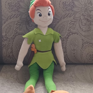 Pode incluir: Boneca de pelúcia do Peter Pan, com túnica, leggings e chapéu verdes, com cabelo e sapatos laranja. A boneca tem um cinto castanho e uma pequena adaga. A boneca tem aproximadamente 46 cm de altura.