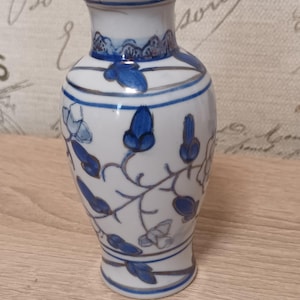 Op de afbeelding: Een witte keramische vaas met een handgeschilderd blauw bloemmotief van ranken en bladeren. De vaas heeft een uitlopende rand, een smalle hals en een rond lichaam versierd met dunne zilverkleurige lijnen die de blauwe botanische motieven omlijnen.