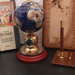 Könnte beinhalten: Ein dekorativer Globus mit tiefblauer Ozeanoberfläche und eingelegten Steinkontinenten, gekrönt von einer goldfarbenen Spitze. Der Globus ruht auf einem goldfarbenen Sockel mit einer runden analogen Anzeige auf einem runden roten Holzständer.