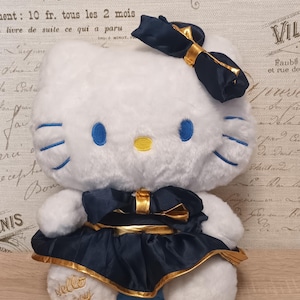 Puede incluir: Un juguete de peluche blanco con forma de gato, ojos bordados en azul, nariz amarilla y bigotes azules. El juguete lleva un vestido azul marino oscuro y un lazo a juego en la cabeza, ambos con detalles dorados. Hello Kitty está bordado en color oro.