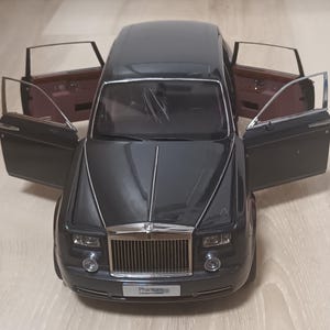 May include: A dark gray scale model of a Rolls-Royce Phantom car with four open doors. The vehicle features a prominent chrome vertical grille, rectangular headlights, and a license plate that reads Phantom. The interior shows brown door panels.