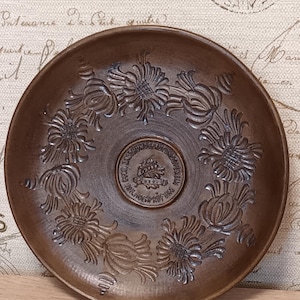 Op de afbeelding: Een ronde bruine keramische schaal met een centraal reliëf-embleem met Duitse tekst en een geweerafbeelding, omringd door een decoratieve ring van acht gestileerde bloemmotieven. De schaal heeft een gladde, matte afwerking en een opstaande rand.