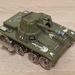 Pode incluir: Um tanque de brinquedo vintage de folha de flandres verde-oliva com uma chave de corda na lateral. O tanque apresenta detalhes impressos, incluindo uma pá, rebites e o texto GAMA TANK. Possui rodas prateadas e uma torre central.