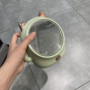 May include: A round, light green handheld mirror featuring two metallic rose gold cat ears at the top and a small, circular rose gold paw print emblem at the bottom. The mirror has a smooth, matte finish and a reflective glass surface.