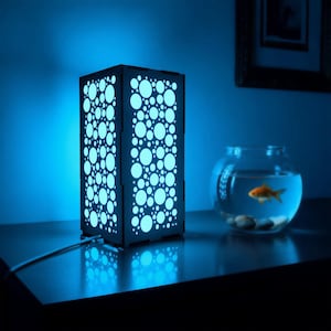 May include: A rectangular wooden lamp with a pattern of varying sized circles, illuminated with a soft blue light. A clear glass fishbowl with a goldfish and small stones sits nearby. The scene is set against a dark blue backdrop.