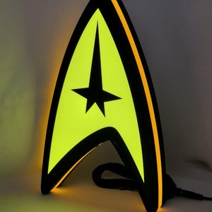 Peut inclure: Une lampe en forme de delta Star Trek avec un centre lumineux jaune vif et un emblème d'étoile noire. Le cadre est noir avec une bordure latérale orange lumineuse, et un cordon d'alimentation noir sort de la base de la structure verticale.