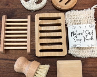 May include: A collection of bath accessories including a round loofah, a round bamboo soap dish, a rectangular bamboo soap dish, a wooden slatted soap rack, a natural sisal soap pouch with a label, a wooden scrub brush, and a square bamboo tray.
