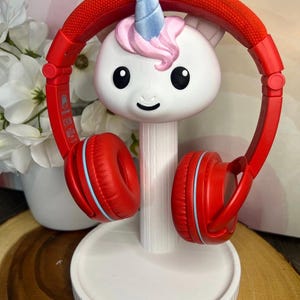 May include: A white unicorn-shaped headphone stand with a blue horn and pink hair, supporting a pair of bright red over-ear headphones. The stand features a cylindrical neck and a circular base, providing a stable structure for the red headset.