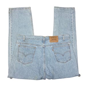 Puede incluir: Vaqueros de mezclilla azul claro con un diseño clásico de cinco bolsillos, un parche de cuero marrón en la cintura trasera y costuras arqueadas características en los bolsillos traseros. El pantalón tiene un corte recto y un lavado claro.