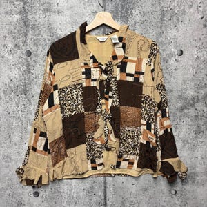 May include: A long-sleeve button-up shirt featuring a patchwork design in shades of brown, tan, and black. The garment includes various patterns such as leopard print, geometric blocks, and abstract line work, with ruffled collar and sleeve cuffs.