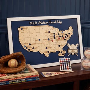 May include: A framed wooden map of the United States titled MLB Stadium Travel Map, featuring cutouts for stadium locations. Includes a baseball glove with a ball, a stack of 1985 World Series Review books, a team logo chart, and a pennant.