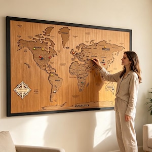 May include: A large rectangular world map wall art piece with a light brown wood grain finish and a black frame. The map features laser-cut continents, country names, a decorative compass rose in the bottom left corner, and colorful push pins.