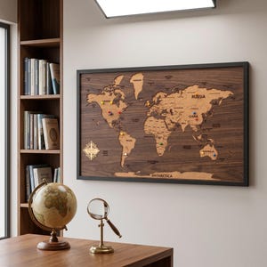 Puede incluir: Un mapa del mundo rectangular para pared con continentes de madera cortados a láser sobre un fondo de veta de madera oscura, enmarcado en negro. El mapa incluye países, océanos y una rosa de los vientos, con chinchetas de colores.