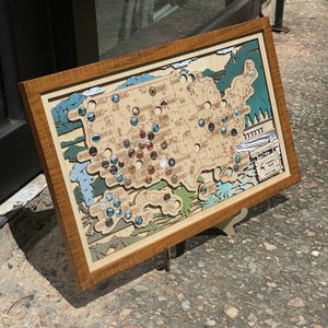 May include: A framed wooden map of the USA, featuring multi-coloured pins. The map is layered with a light brown wood base, and the frame is a darker brown. The map includes the text "Travel Map" and "Lakes & Lands".