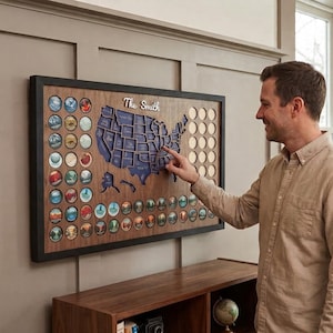 Può includere: Una mappa da parete rettangolare in legno degli Stati Uniti con cornice scura, sagoma della mappa blu e la scritta The Smith. La tavola include fessure circolari per raccogliere gettoni, con molti inserti circolari colorati raffiguranti paesaggi.