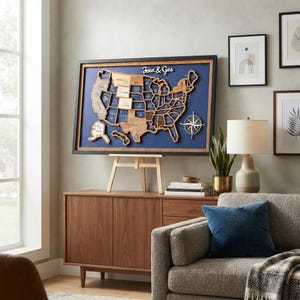 May include: A framed wooden map of the United States featuring a dark blue background, a compass rose, and the text "Jenn & Gus" in white script. The map is composed of individual wooden state pieces, some engraved with text, mounted on a wooden easel.