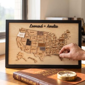 Puede incluir: Un mapa de madera enmarcado de los Estados Unidos con piezas de estados cortadas con láser en varios tonos de madera. Los nombres Leonard + Amelia aparecen en letra cursiva negra en la parte superior. Incluye Alaska y Hawái.