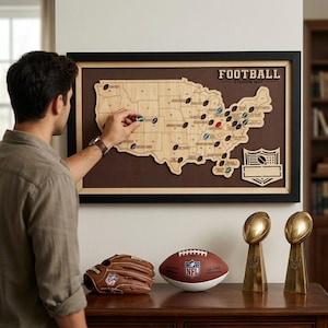 May include: A framed wooden map of the United States titled FOOTBALL STADIUM MAP, featuring small football-shaped markers placed across the map. Below, a brown leather baseball glove, a football, and two gold-colored trophy statues are displayed.