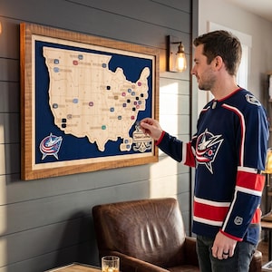 Puede incluir: Un mapa de pared de madera de los Estados Unidos con un fondo de fieltro azul oscuro, un acabado de madera clara y varios marcadores pequeños de colores colocados en los estados. El mapa incluye un logotipo y el texto TimberCraftMap.
