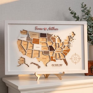 May include: A framed wooden USA travel map featuring layered states in varying shades of brown and tan. The map includes a compass rose, the text "Fiona ♥ Arthur" at the top, and "USA TRAVEL MAP ADVENTURE AWAITS" at the bottom, all set in a white frame.