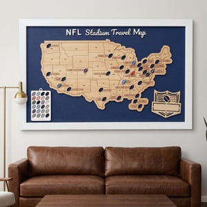 May include: A framed wall map of the United States featuring a light wood cutout of the country on a dark blue background. The map includes the text NFL Stadium Travel Map, stadium locations marked with small football icons, and a separate legend.