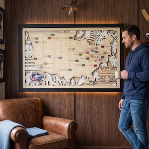 Könnte beinhalten: Ein großes rechteckiges Wandkunstwerk aus Holz mit einer mehrschichtigen Karte der Vereinigten Staaten und Eishockey-Motiven, darunter eine Torwartmaske, Eishockeyspieler und der Schriftzug TimberCraftMap, schwarz gerahmt mit Hintergrundbeleuchtung.