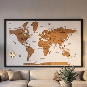 Może przedstawiać: Oprawiona mapa świata z białym tłem i brązowymi kontynentami z korka. Mapa zawiera czarne etykiety tekstowe dla krajów i oceanów, logo róży wiatrów oraz kilka kolorowych pinezek wbitych w różne lokalizacje na całym globie.