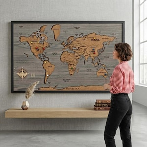 Pode incluir: Um grande mapa-múndi retangular montado na parede com continentes de madeira cortados a laser em um acabamento marrom natural sobre um fundo de madeira cinza. O mapa inclui etiquetas pretas para países e oceanos, com percevejos coloridos.