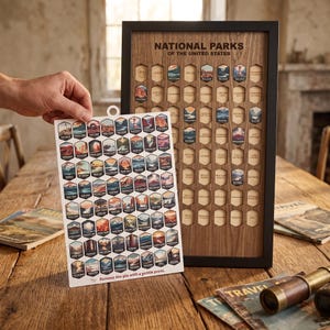 May include: A wooden National Parks of the United States tracker board in a black frame, featuring hexagonal cutouts for park tokens. A white sheet with colorful hexagonal stickers of various national parks is held up in front of the board.