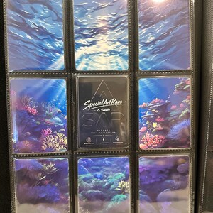 May include: A black trading card binder page containing nine rectangular cards. The top row shows blue water surfaces. The middle row features coral reefs and a central black card with the text Special Art Rare, ASAR, SAP, and social media icons.
