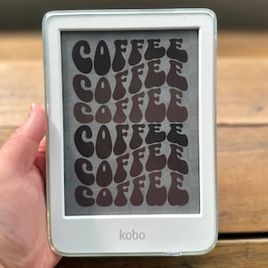 May include: A white Kobo e-reader featuring a screen display with the word COFFEE repeated in a wavy, dark brown, retro-style font. The device has a clear protective case and a rectangular frame with the Kobo logo printed at the bottom center.