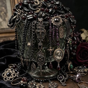 May include: A collection of dark-toned jewelry including beaded necklaces, ornate metal pendants with cross and cameo motifs, and various rings with dark gemstones, all draped over and around a clear glass pedestal bowl.