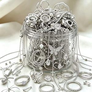 May include: A clear plastic jar filled with an assortment of silver-toned jewelry, including rings, necklaces, and chains. The collection features various charms such as butterflies, crosses, and teardrop-shaped crystals, with some pieces spilling out.