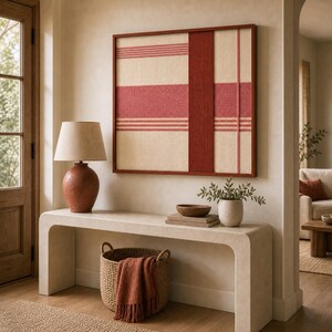 May include: A framed wall art piece with a woven design in cream and red hues. The artwork hangs above a console table with a lamp, a basket, and decorative items. The room has a neutral color palette.