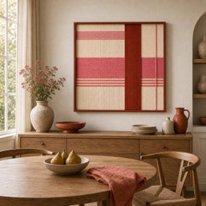 May include: A framed wall art piece with a woven design in shades of red and beige. The artwork features horizontal stripes and a vertical panel, creating a geometric pattern. The frame is a dark reddish-brown color. The scene includes a wooden table and cabinet.