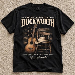 May include: A black short-sleeve t-shirt featuring a graphic print of an acoustic guitar, a cowboy hat resting on a wooden chair, and a distressed American flag. The text reads BRIAN 'NASHVILLE' DUCKWORTH, COUNTRY MUSIC STAR, and Brian Duckworth.