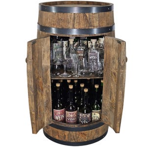 Può includere: Un mobile bar a forma di botte in legno marrone con due ante a battente e fasce in metallo nero. L'interno presenta due ripiani contenenti varie bottiglie di vetro, tra cui quelle etichettate Whisky, Wein e Vino, insieme a diversi bicchieri.