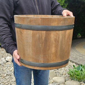 Peut inclure: Un pot de fleurs cylindrique en bois avec une finition naturelle aux tons brun clair. Le pot est renforcé par deux bandes métalliques horizontales gris foncé, chacune fixée par des rivets métalliques visibles, offrant une structure robuste.