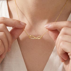 May include: A gold-toned infinity symbol necklace featuring a delicate chain and a central pendant encrusted with small, clear, sparkling stones along the curved surface of the infinity loop, creating a refined and polished appearance.
