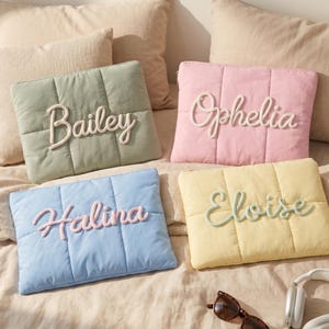 May include: Four rectangular quilted pouches in sage green, pastel pink, light blue, and pale yellow. Each pouch features a raised, cursive name appliqué: Bailey in white, Ophelia in white, Halina in pink, and Eloise in sage green.