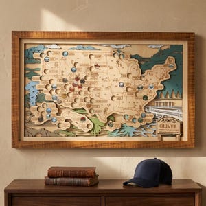 May include: A framed, multi-layered wooden map of the United States featuring cutouts for national parks, engraved state borders, and circular tokens. A sign in the lower right corner reads OLIVER NATIONAL PARK ADVENTURES. A navy blue baseball cap sits below.