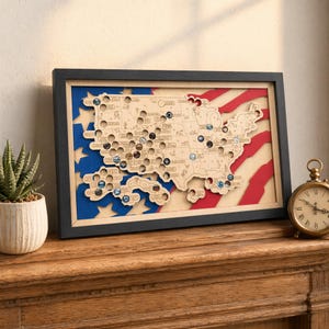May include: A rectangular framed wall art piece featuring a laser-cut wooden map of the United States layered over a blue and red flag design. The map includes circular cutouts for tokens, with several colorful circular tokens placed inside.