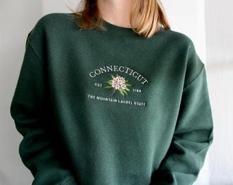 May include: A dark forest green crewneck sweatshirt featuring white embroidered text that reads CONNECTICUT EST 1788 THE MOUNTAIN LAUREL STATE, centered with a detailed embroidered illustration of pink mountain laurel flowers and green leaves.