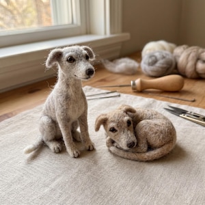 May include: Two needle-felted dog figurines made from light gray and beige wool fibers. One dog is sitting upright with alert ears, while the other is curled into a sleeping position. Both figures feature dark eyes and noses, showcasing a textured finish.