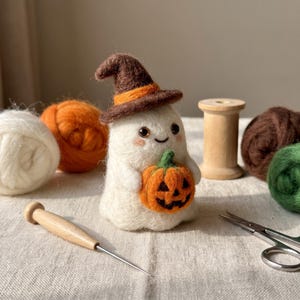 Puede incluir: Una figura de fantasma blanco de lana afieltrada con un sombrero de bruja marrón con banda naranja, sosteniendo una pequeña calabaza naranja. El fantasma tiene ojos de cuentas negras y mejillas rosadas. Hay ovillos de lana y una herramienta.