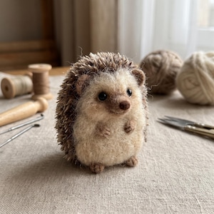 May include: A needle-felted hedgehog figurine with a cream-colored face and belly, featuring dark brown, spiky textured quills on its back, small black eyes, and a brown nose, standing upright with tiny paws and feet visible on its front.