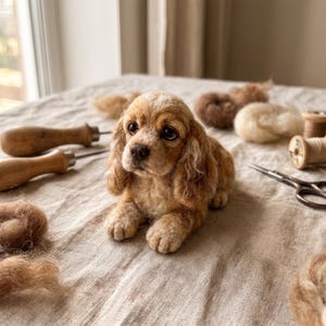 May include: A needle-felted sculpture of a light brown and cream-colored spaniel puppy with long, wavy ears and dark eyes. The puppy is positioned among crafting supplies, including two wooden-handled felting tools, metal scissors, and spools.