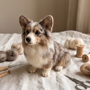 Può includere: Una statuetta di Corgi in lana infeltrita con un mantello maculato nei toni del grigio, marrone e bianco. Il cane ha orecchie dritte, occhi scuri e il petto bianco. È circondato da lana cardata, strumenti in legno e forbici in metallo.